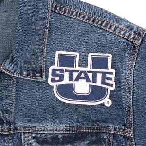 May include: Close-up of a denim jacket with a patch. The patch is white with navy blue lettering that reads "STATE" and a stylized "U" shape. The jacket is a medium wash denim with visible stitching.