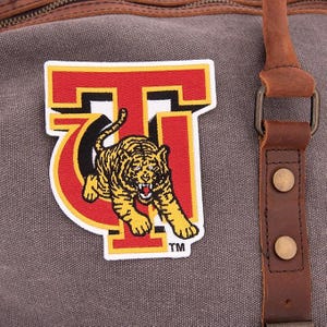 May include: A gray canvas duffel bag with a large patch featuring a tiger graphic. The patch is in red, yellow, and black, with the letters "T" and "G" incorporated into the design. Brown leather straps and accents are visible.