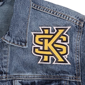 May include: Close-up of a blue denim jacket with a yellow and white embroidered patch. The patch features the letters "SK" in a stylized design. The jacket has visible stitching and a classic collar.