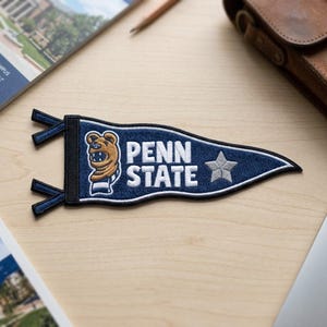 May include: A navy blue Penn State pennant patch with white embroidered text and a star. The patch features a brown bear mascot and is trimmed with white stitching. The pennant has a black border with navy blue ties.