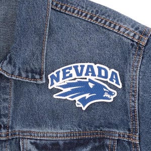 May include: Close-up of a blue denim jacket with a stitched design. The jacket features a white and blue patch with the word "NEVADA" and a wolf logo. The jacket has brown stitching.