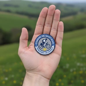 Peut inclure: Un écusson brodé circulaire avec une bordure bleue portant le texte Camino de Santiago et Peregrino on Wheels. Le centre présente un fond gris avec la silhouette noire d'un cycliste et une petite icône de coquille Saint-Jacques jaune.