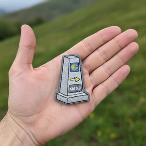 Peut inclure: Un écusson brodé gris en forme de borne kilométrique. Le motif présente un carré bleu avec une icône de coquille Saint-Jacques jaune, une flèche jaune pointant vers la droite et un rectangle contenant le texte noir KM 0.0 sur fond gris.