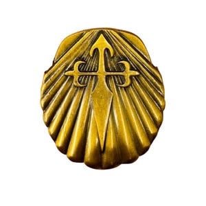 May include: A gold-toned metal pin shaped like a scallop shell with a raised relief of a cross of Saint James in the center. The shell features radiating ridges extending from the base to the outer edges, creating a textured, metallic surface.