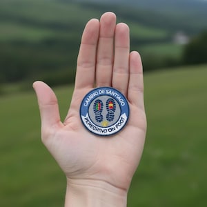 Op de afbeelding: Een ronde geborduurde patch met een grijs midden en twee donkerblauwe voetafdrukken van laarzen, elk met kleine rode, groene en gele accenten. De rand is donkerblauw met de witte tekst CAMINO DE SANTIAGO PEREGRINO ON FOOT en een gele schelp.