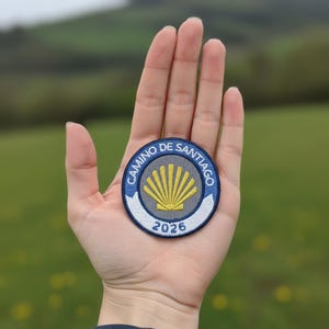 May include: A circular embroidered patch featuring a yellow scallop shell on a gray background, surrounded by a blue border with the text CAMINO DE SANTIAGO in white, and a white banner at the bottom displaying the year 2026 in blue embroidery.