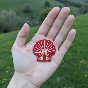 Könnte beinhalten: Ein rot-cremefarbener gestickter Aufnäher in Form einer Jakobsmuschel. Das Design zeigt die zentrale Silhouette einer Person mit einem Wanderstock, umgeben von strahlenförmigen cremefarbenen Linien auf einem einfarbig roten Hintergrund.