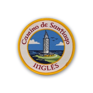 May include: A round embroidered patch featuring a lighthouse on a green hill with blue water and sky. The patch has the words "Camino de Santiago" and "Ingles" in red text, with a yellow border.