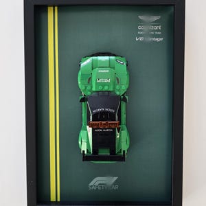 Op de afbeelding: Ingelijste kunstwerk met een groene Aston Martin Lego-auto. De auto staat tegen een donkergroene achtergrond met gele strepen en de woorden "SAFETY CAR". De lijst is zwart en het kunstwerk bevat het logo van het Cognizant Formula One Team.