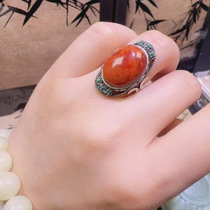 以下が含まれることがあります： 大きな楕円形の赤みがかったオレンジ色の宝石をあしらったシルバーリング。リングは小さな緑色の石で装飾されています。リングは人の指にあります。