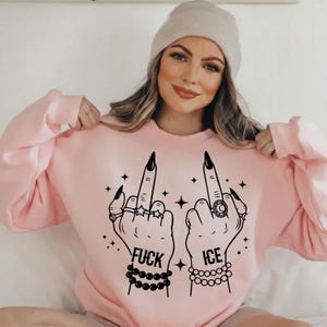 May include: A light pink sweatshirt with a graphic of two hands giving the middle finger. The hands are adorned with rings and bracelets, and the text "FUCK ICE" is printed below. The sweatshirt has long sleeves and a crew neck.