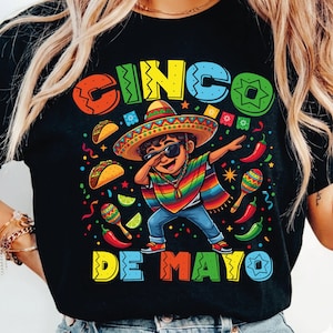 May include: Black t-shirt featuring a colorful Cinco de Mayo design. The graphic includes a cartoon boy in a sombrero and sunglasses, dabbing. The words "CINCO DE MAYO" are displayed in vibrant colors, along with festive illustrations.