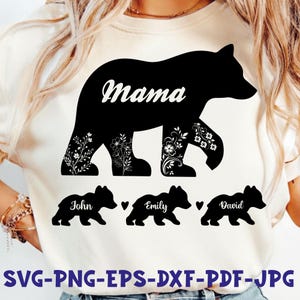 Może przedstawiać: Kremowy t-shirt z czarną sylwetką niedźwiedzia i białym napisem "Mama". Poniżej znajdują się trzy mniejsze sylwetki niedźwiedzi z imionami "John", "Emily" i "David". Kwiatowe wzory zdobią nogi niedźwiedzia. Na dole koszulki napis "SVG-PNG-EPS-DXF-PDF-JPG".