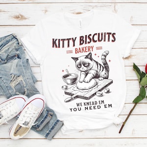 May include: White t-shirt with a graphic of a grumpy cat kneading dough on a wooden board. Text above reads "KITTY BISCUITS BAKERY" and below "WE KNEAD EM YOU NEED EM". The shirt is paired with blue jeans and white sneakers.