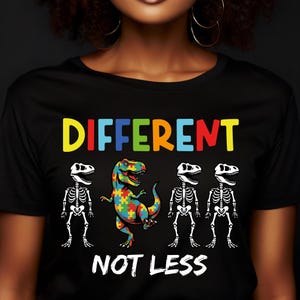 Könnte beinhalten: Schwarzes T-Shirt mit den Worten "DIFFERENT NOT LESS" in bunten Buchstaben. Das Shirt zeigt einen Tyrannosaurus Rex mit einem Puzzlemuster und drei Skelett-Dinosauriern.