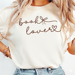 Puede incluir: Camiseta color crema con las palabras "book lover" en escritura marrón. La palabra "book" tiene un lazo, y "lover" tiene un corazón. La camiseta es de estilo casual, con mangas cortas.