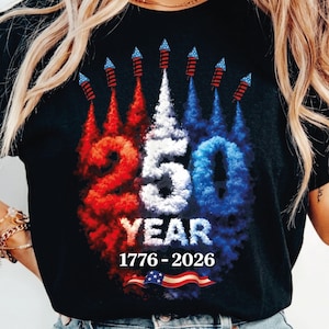 Pode incluir: Camiseta preta com um design patriótico. O design apresenta o número "250" em branco, vermelho e azul, com "YEAR" abaixo e as datas "1776-2026". Fogos de artifício e uma bandeira americana também estão incluídos.