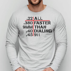 May include: A light gray long-sleeve shirt with black text and a red crosshair design. The text reads ".22 ALL .380 FASTER 9MM THAN .40 DIALING .45 911". The shirt is made of a soft material.