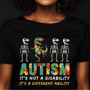May include: Black t-shirt with a colorful graphic featuring a dinosaur made of puzzle pieces, flanked by skeleton dinosaurs. The word "AUTISM" is displayed in puzzle piece letters, with the text "IT'S NOT A DISABILITY IT'S A DIFFERENT ABILITY" below.