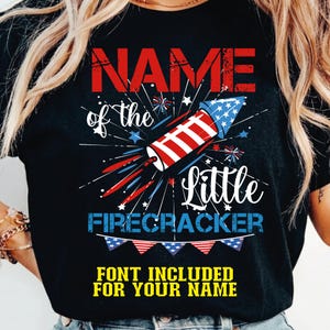 Puede incluir: Camiseta negra con un diseño de fuegos artificiales en rojo, blanco y azul. El texto dice "NAME of the Little FIRECRACKER" y "FONT INCLUDED FOR YOUR NAME". El diseño es patriótico.