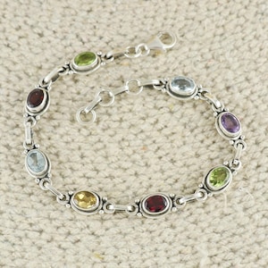 May include: A silver bracelet featuring oval-shaped gemstones in various colors, including green, red, yellow, blue, and purple. The bracelet has a secure clasp and is displayed on a neutral-toned surface. This jewelry piece is suitable for everyday wear or special occasions.