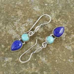 May include: A pair of silver dangle earrings featuring a teardrop-shaped, deep blue stone, a round turquoise stone, and a silver leaf accent. The earrings are displayed on a textured, neutral-toned surface.