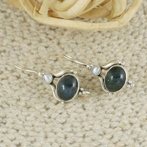 Puede incluir: Un par de pendientes de plata con piedras preciosas ovaladas de color azul verdoso oscuro. Cada pendiente tiene una pequeña piedra de acento redonda blanca y un diseño plateado decorativo. Los pendientes se muestran sobre una superficie beige texturizada.