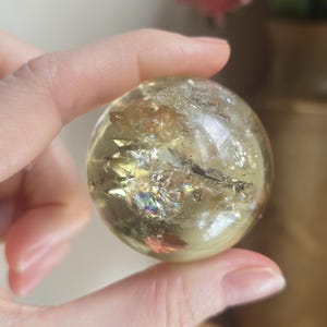 May include: A clear, spherical crystal ball with internal inclusions and iridescent flashes. The sphere is held between fingers, showcasing its translucent quality and the play of light within. The crystal has a yellow-gold hue.