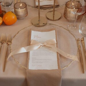 May include: Elegant table setting with gold-toned cutlery, glassware, and candle holders. A menu card is tied with a cream ribbon, and a napkin is placed on a clear charger plate. An orange fruit is visible.