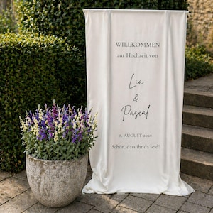 Könnte beinhalten: Ein vertikales Banner aus cremeweißem Stoff mit dem schwarzen Aufdruck WILLKOMMEN zur Hochzeit von Lia & Pascal 8. AUGUST 2026 Schön, dass ihr da seid! Das Banner hängt an einer horizontalen Stange und fällt in weichen Falten herab.
