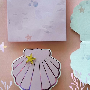 Puede incluir: Una manualidad de papel en forma de concha de color rosa claro con un adorno de estrella de mar amarilla. La concha tiene acentos iridiscentes y se encuentra sobre un fondo rosa con ilustraciones acuáticas, incluyendo peces y burbujas. También hay un bloc de notas cuadrado con ilustraciones acuáticas similares.