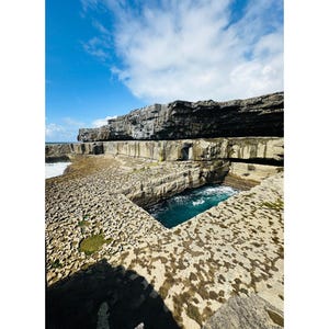 May include: A coastal landscape with a layered rock formation and a turquoise pool. The sky is blue with clouds. The image includes the text "POLL NA BPÉIST" and "INIS MÓR".