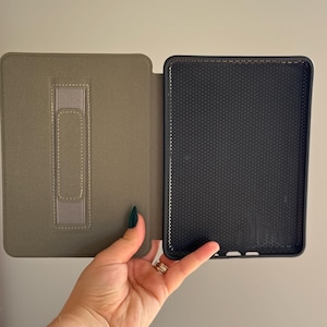 May include: A protective e-reader case featuring a sage green exterior with a vertical hand strap on the inner cover and a black interior shell with a textured honeycomb pattern designed to hold a digital reading device securely in place.