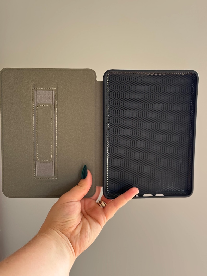 May include: A protective e-reader case featuring a sage green exterior with a vertical hand strap on the inner cover and a black interior shell with a textured honeycomb pattern designed to hold a digital reading device securely in place.