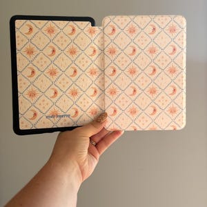 May include: A black e-reader case with a light cream interior featuring a repeating diamond pattern of pink suns and crescent moons. The interior surface includes the text STAY PRETTY printed in a small blue font near the bottom left corner.