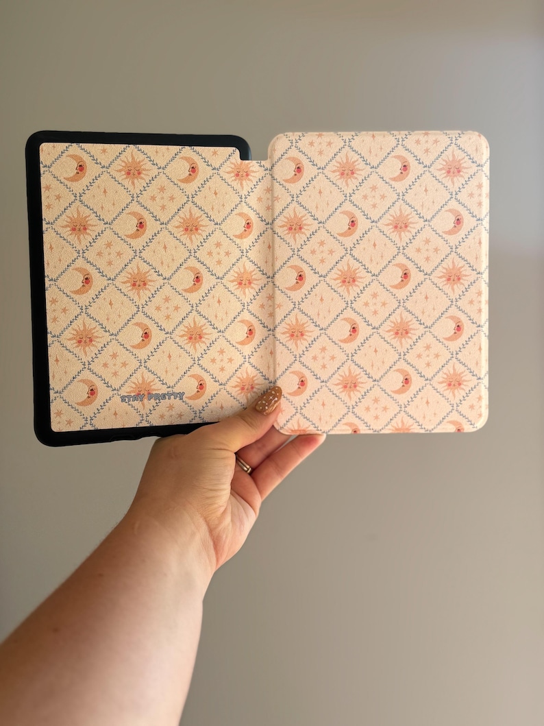 May include: A black e-reader case with a light cream interior featuring a repeating diamond pattern of pink suns and crescent moons. The interior surface includes the text STAY PRETTY printed in a small blue font near the bottom left corner.