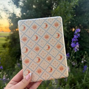 May include: A rectangular e-reader case featuring a cream-coloured background with a repeating diamond grid pattern outlined in blue vines. The design includes alternating illustrations of orange suns with faces and crescent moons with faces, plus small stars.