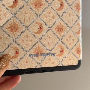 May include: A tablet case featuring a repeating diamond pattern outlined in blue vines. The design includes alternating sun and crescent moon faces in shades of peach and orange, with small stars. The text STAY PRETTY is printed in blue.