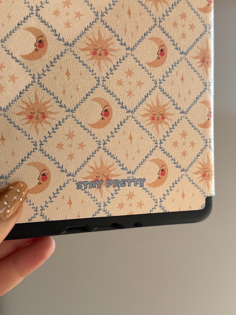 May include: A tablet case featuring a repeating diamond pattern outlined in blue vines. The design includes alternating sun and crescent moon faces in shades of peach and orange, with small stars. The text STAY PRETTY is printed in blue.