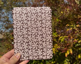 以下が含まれることがあります： 長方形のノートカバー。花柄のデザインで、明るいピンクの花が濃い茶色の背景に繰り返されています。ノートは、緑と茶色の葉のぼやけた背景の前に保持されています。