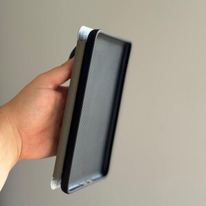 May include: A rectangular protective case for a mobile device featuring a black frame with a textured interior surface and a solid grey exterior panel. The case has a slim profile and a smooth finish, designed to fit around the edges of a phone.