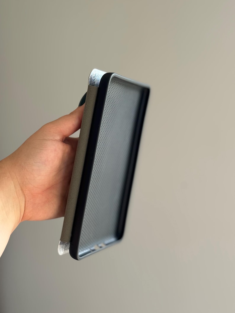 May include: A rectangular protective case for a mobile device featuring a black frame with a textured interior surface and a solid grey exterior panel. The case has a slim profile and a smooth finish, designed to fit around the edges of a phone.