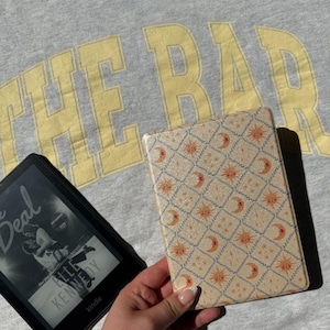 May include: A black e-reader displaying the book cover for The Deal by Elle Kennedy next to a rectangular e-reader case featuring a cream-coloured diamond grid pattern with orange suns and crescent moons, set against a grey shirt with yellow text.