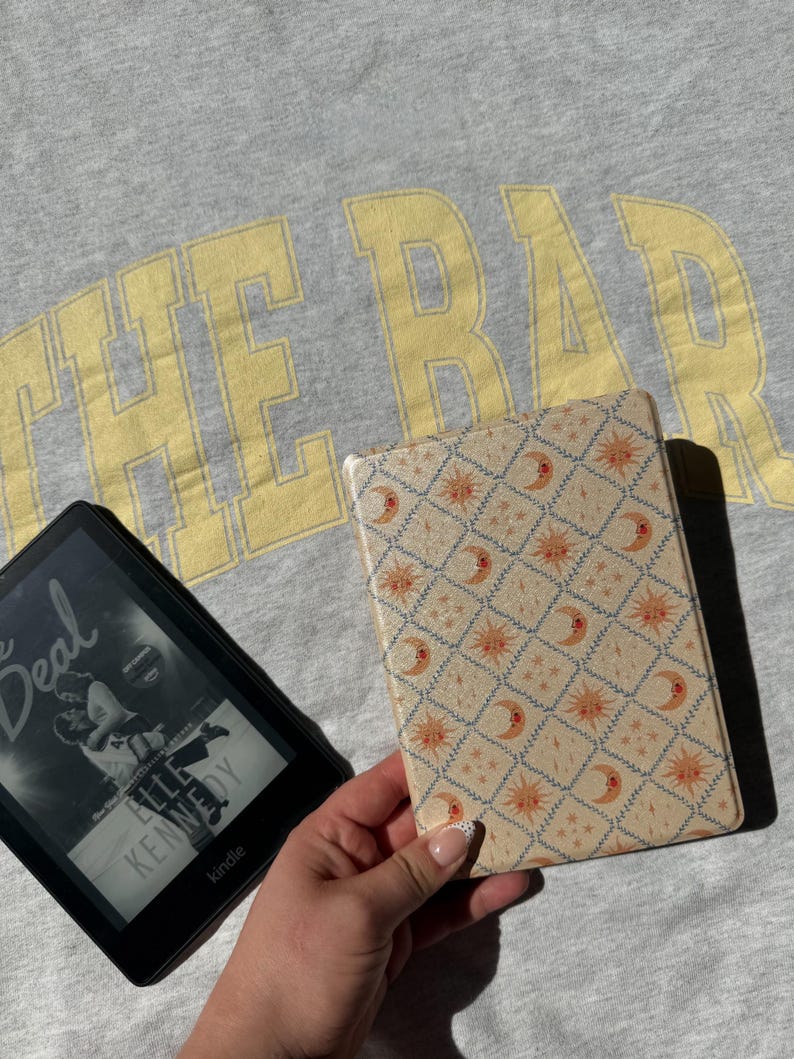 May include: A black e-reader displaying the book cover for The Deal by Elle Kennedy next to a rectangular e-reader case featuring a cream-coloured diamond grid pattern with orange suns and crescent moons, set against a grey shirt with yellow text.