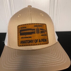 Op de afbeelding: Tan en witte truckerpet met een leren patch. De patch toont een kogeldiagram met labels en de tekst "Anatomy of a Pew". De pet heeft een gebogen klep en een mesh achterkant.