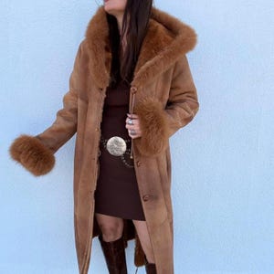 May include: A long, brown suede coat with a fur-lined hood and cuffs. The coat has a button-down front and a belt with a silver buckle. The model is wearing a brown dress and boots. The coat is a vintage style.