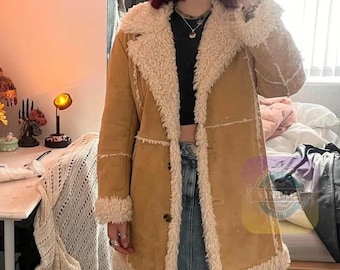 May include: A tan suede coat with a cream-colored shearling lining and trim. The coat has a button closure and a classic, longline silhouette. The shearling trim is visible on the collar, cuffs, and along the front edges.