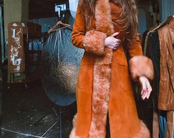 Può includere: Un lungo cappotto in camoscio color ruggine con finiture in ecopelliccia sul colletto, sui polsini e sull'orlo. Il cappotto ha un design classico con due tasche anteriori. La finitura in pelliccia è di una tonalità marrone più chiara.