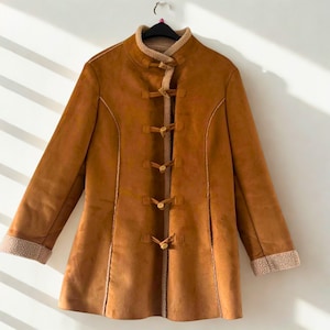 May include: A brown suede-like coat with a high collar and toggle closures. The coat has a soft, beige lining visible at the cuffs and collar. The coat is hanging on a black hanger against a white wall.