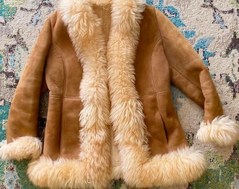 Può includere: Giacca in camoscio marrone con finiture in ecopelliccia color crema. La giacca presenta un ampio colletto, polsini e un bordo anteriore in pelliccia di peluche. La giacca è aperta, mostrando la fodera interna.